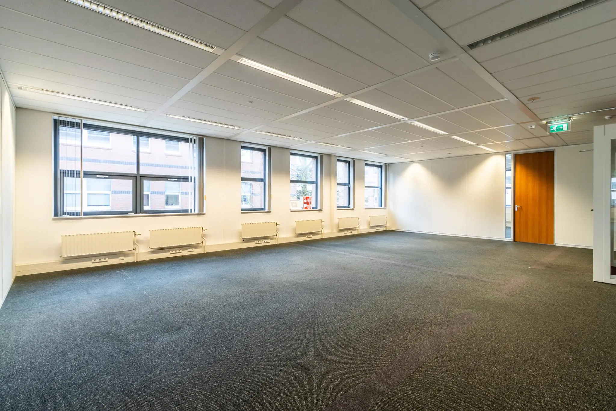 Leegstaand, licht kantoorruimte aan de Lange Dreef met meerdere ramen, radiatoren en een gesloten houten deur.